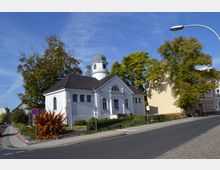 Weißes Gebäude mit klassizistischer Architektur, einschließlich Säulen und einem Rundbogenfenster über dem Eingang, sowie einem kleinen Turm mit Kuppeldach. Das Gebäude ist von Bäumen und gepflegten Grünflächen umgeben und liegt an einer gepflasterten Straße.