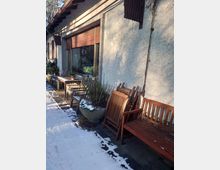 Außenbereich eines Gebäudes mit weißer Putzfassade, großen Fenstern und geschlossenen Rollläden. Vor dem Gebäude stehen Holzbänke, Tische und Stühle, teils in Schnee bedeckt; daneben eine große Pflanzschale.