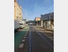 Straßenansicht mit Straßenbahngleisen, Fahrradweg und Gehweg in einer urbanen Umgebung. Links stehen mehrere historische Gebäude und geparkte Fahrzeuge, rechts befinden sich moderne Gebäude, eine überdachte Bushaltestelle und eine erhöhte grüne Brücke.