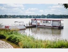 Ein weiß-rotes Ausflugsboot mit der Aufschrift „Mein Dampfer“ liegt an einem kleinen Steg in einem ruhigen See, umgeben von Ufervegetation. Im Hintergrund sind Bäume und vereinzelte Gebäude zu sehen, unter einem Himmel mit weißen Wolken.