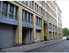 Mehrstöckiges Verwaltungsgebäude mit beigefarbener Fassade und großen Glasfenstern, flankiert von beschilderten Ein- und Ausfahrten einer Tiefgarage. Es befindet sich an einer schmalen, gepflasterten Straße in städtischer Umgebung.