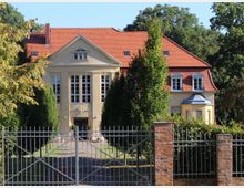 Gelbes Herrenhaus mit rotem Satteldach und großen Fenstern, umgeben von gepflegten Bäumen und einer gepflasterten Auffahrt, die hinter einem schmiedeeisernen Tor mit Backsteinsäulen zum Eingang führt. Im Hintergrund sind hohe Bäume zu sehen.