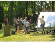 Filmset in einem grünen Park mit Bäumen, mehreren Personen und technischem Equipment wie Kameras, Stativen und einem großen Lichtreflektor. Im Vordergrund ist ein Grabstein zu sehen, was auf einen Friedhof als Drehort hindeuten könnte.
