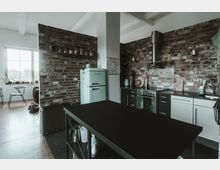 Moderne Küche mit Backsteinwänden, schwarzer Arbeitsplatte und Edelstahlgeräten. Neben einem mintgrünen Retro-Kühlschrank befindet sich eine Küchenzeile mit integriertem Herd und Dunstabzugshaube, im Hintergrund ein Essbereich mit Tisch und Stühlen.