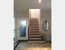 Treppenhaus in einem modernen Wohnhaus mit einer Holztreppe und Metallhandlauf, die zu einer oberen Etage führt. Links eine Glastür mit mattem Muster, rechts ein ovaler Spiegel und dekorative Laternen auf der unteren Treppenstufe; der Boden ist mit beigefarbenen Fliesen ausgelegt.