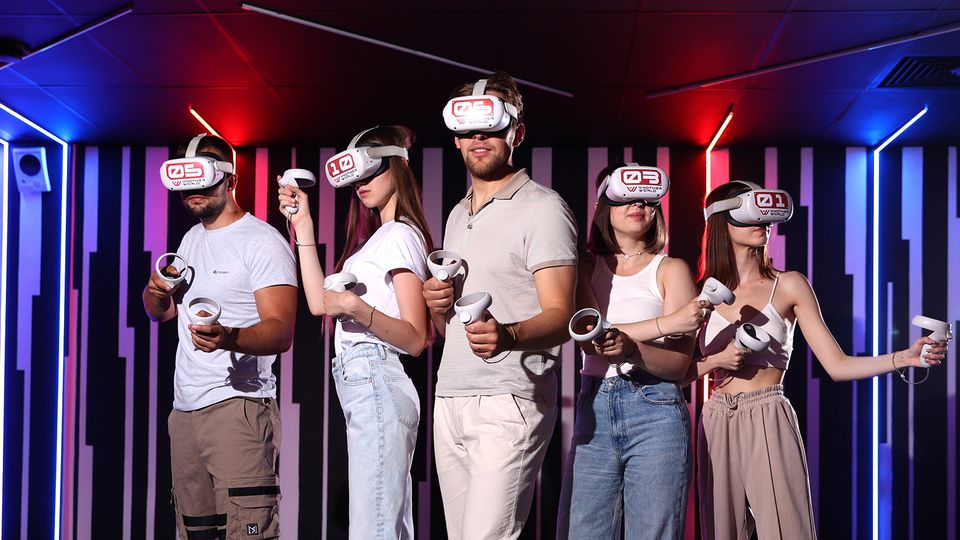 Innenraum einer VR-Arcade mit dunkler Decke, LED-Lichtleisten in Rot und Blau und einer Wand aus vertikalen Paneelen. Mehrere VR-Headsets und Hand-Controller sind im Vordergrund zu sehen, was den Raum als Virtual-Reality-Spielbereich kennzeichnet.