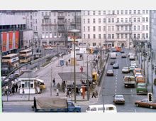 Städtischer Platz mit einer U-Bahn-Station, umgeben von mehrstöckigen Altbauten. Auf der rechten Seite verläuft eine belebte Straße mit parkenden und fahrenden Autos, während links Busse und Fußgänger zu sehen sind. Im Zentrum des Bildes steht ein Denkmal.