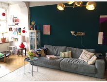 Wohnzimmer mit einer petrolfarbenen Wand, grauem Sofa und dekorativen Kissen. Das Zimmer ist mit einem weißen Teppich, einer Vitrine, einem roten Lampensockel und Bücherregalen ausgestattet; helle Holzdielen und ein Tisch mit Tulpen ergänzen den Raum.