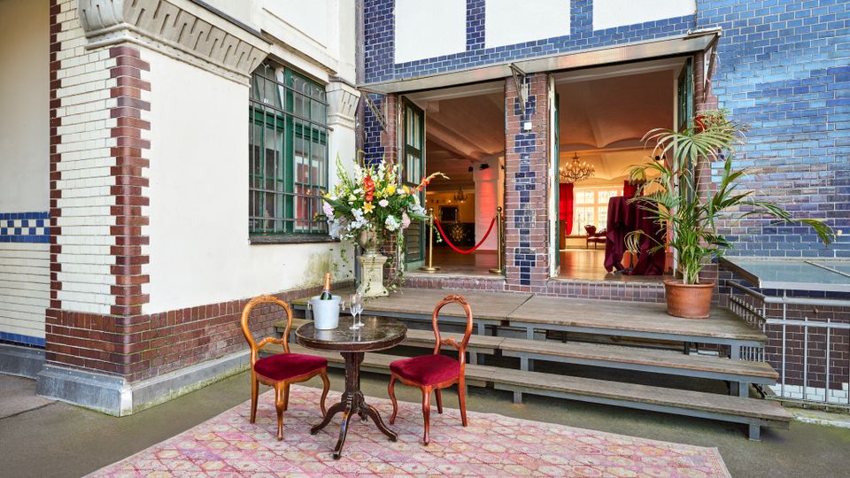 Eingangsbereich eines Gebäudes mit blau-weiß gekachelter und verputzter Fassade, großen Fenstern und einer offenen Tür zu einem Innenraum mit Kronleuchtern. Davor stehen auf einem gemusterten Teppich ein runder Tisch mit zwei roten Polsterstühlen, daneben Blumendekor, Topfpflanze und eine kleine Treppe zum Eingang.