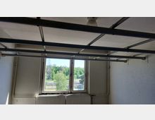 Ein leerer Raum mit weiß gestrichenen Wänden und einer Metallkonstruktion unter der Decke. Im Hintergrund befindet sich ein Fenster mit Blick auf grüne Bäume, einen Strommast und einen Zaun, darunter ein Heizkörper.