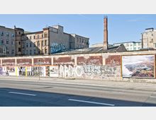 Straßenszene mit einer alten, teilweise verfallenen Backsteinfassade im Hintergrund und einer betonierten, mit Graffiti versehenen Mauer im Vordergrund, die mit Plakaten beklebt ist. Ein hoher Schornstein und mehrgeschossige Gebäude deuten auf ein ehemaliges Industrie- oder Wohngebiet hin.