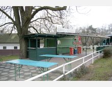 Eine einfache Grillstation mit grün gestrichener Holzverkleidung und Flachdach, umgeben von Bäumen. Vor der Station stehen Biertischgarnituren und rote Coca-Cola-Tonnen stehen als Dekoration oder Abfalleimer bereit.