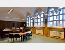 Innenansicht eines historischen Lesesaals mit hohen, gewölbten Decken und großen Fenstern, die in mehrere kleine Felder unterteilt sind. Holzwände, Heizkörper unter den Fenstern und ein Tisch mit Stühlen prägen die Einrichtung, ergänzt durch Schreibtische und Deckenlampen.