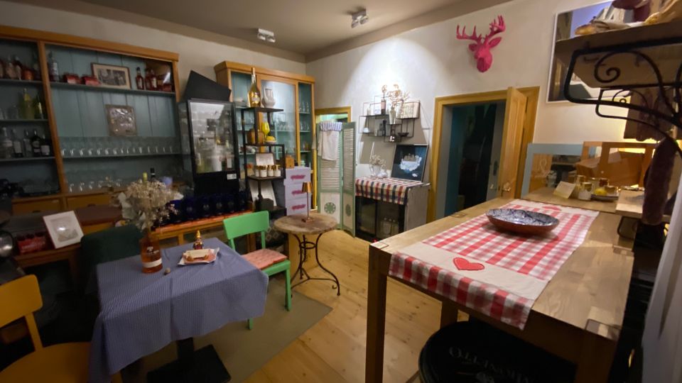 Gemütlicher Innenraum eines kleinen Cafés oder Ladens mit Holzfußboden, Vitrinen und Regalen voller Flaschen und Gläser sowie einem Tisch mit blauem Tischtuch und bunten Stühlen. Im Vordergrund steht eine hohe Holztheke mit rot-weiß kariertem Läufer, an der Wand hängen Dekorationen und Regalbretter mit weiteren Produkten.