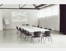 Ein heller Seminarraum mit weiß gestrichenen Wänden, einer langen, rechteckigen Tafel, die von schwarzen Stühlen umgeben ist. Im Hintergrund befinden sich eine Leinwand mit einem projizierten Bild, ein Flipchart und große, bodenlange weiße Vorhänge vor den Fenstern.