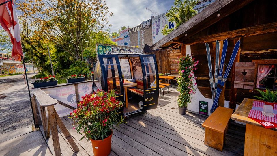 Holzterrasse eines rustikalen Lokals mit Holzbänken und Tischen, bepflanzten Kübeln und einer dekorativen Gondelkabine als Sitzbereich. Daneben eine hölzerne Hütte mit an der Wand befestigten Skiern; im Hintergrund Bäume sowie Gebäude mit Graffiti und Baustellenkran.