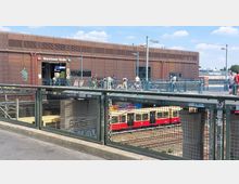 Bahnhof Warschauer Straße in Berlin, mit einer braunen Fassade und mehreren Personen auf der oberen Plattform. Im unteren Bereich sind Gleise sowie ein gelb-roter S-Bahn-Zug zu sehen, im Vordergrund ein mit Graffiti versehenes Gittergeländer.