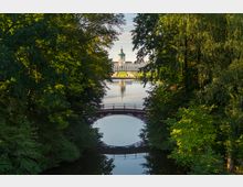 Schloss Charlottenburg in Berlin, gesehen durch eine von Bäumen gesäumte Allee mit einem kleinen Teich und einer gewölbten Brücke im Vordergrund. Das Schloss spiegelt sich im Wasser und ist von gepflegtem Rasen umgeben.