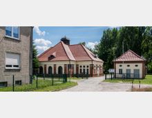 Historisches Gebäudeensemble mit roten Ziegeldächern, bestehend aus einem zentralen Pavillon mit Rundbogenfenstern und einem kleineren Nebengebäude, umgeben von Grünflächen und einem Zaun. Im Hintergrund sind hohe Bäume und ein blauer Himmel sichtbar.