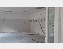 Leerer, gefliester Innenraum eines ehemaligen Industrie- oder Küchenbereichs mit erhöhter Plattform und Wänden aus weißen Fliesen mit blauen Mustern. Abgehängte Decke mit Leuchtstofflampen und eine Glastür mit Verbotsschild am rechten Bildrand.