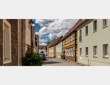 Schmale Kopfsteinpflasterstraße in einer Altstadt, gesäumt von Häusern mit hellen Fassaden, darunter ein Fachwerkhaus in Gelb und Braun. Laternen und vereinzelte Pflanzen geben der Szene ein ländliches Ambiente, während einige parkende Autos sichtbar sind.