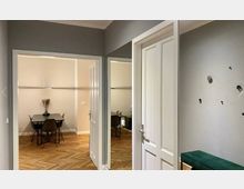 Heller Innenraum in einer Wohnung mit Parkettboden, einer offenen Tür, die in ein Esszimmer mit einem Tisch und vier schwarzen Stühlen führt. Die Wände sind in neutralen Farben gestrichen, und die Einrichtung wirkt minimalistisch.