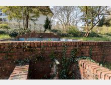 Backsteinmauer mit Bewuchs aus Efeu und Pflanzen umgibt ein leeres, verwittertes Wasserbecken im Freien. Im Hintergrund stehen Bäume, Sträucher und ein Wohnhaus in einer ruhigen Gartenlandschaft.