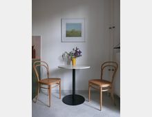 Eine minimalistisch eingerichtete Ecke mit einem runden Tisch mit schwarzem Metallfuß, zwei Holzstühlen mit geflochtenen Sitzflächen sowie einer gelben Vase mit Blumen. An der weißen Wand hängt ein gerahmtes Landschaftsbild, und der Boden ist mit sechseckigen Fliesen ausgelegt.