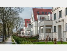 Reihen moderner, mehrstöckiger Wohnhäuser mit weißen Fassaden und roten Ziegeldächern entlang einer baumbestandenen Straße mit Gehweg und gepflegter Grünanlage. Im Vordergrund sind Hecken und kahle Äste sichtbar, die an einem sonnigen Tag Schatten werfen.