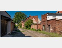 Eine schmale Gasse mit Kopfsteinpflaster, umgeben von alten Backsteingebäuden, teils mit weißen Putzfassaden und Efeu bewachsen. Im Hintergrund steht ein großer Baum, und der Himmel ist klar und blau.
