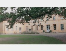 Gelbes historisches Gebäude mit symmetrischer Fassade, mehreren hohen Fenstern, einer verzierten Eingangstür und klar strukturierten Details. Im Vordergrund sind ein Baum, ein gepflegter Garten und eine gepflasterte Fläche sichtbar.