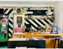 Ein kleiner Laden mit einem Tresen aus Holz, auf dem Blumen, Schmuck und Schallplatten ausgestellt sind. Im Hintergrund sind schwarz-weiß gestreifte Wände, ein Regal mit Gläsern und Getränken sowie eine handgeschriebene Tafel mit der Aufschrift „Getränke“ zu sehen.