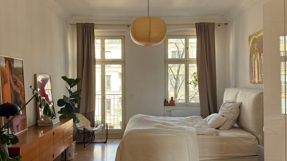 Bright, minimalist bedroom with a wooden floor, a double bed with white bedding, and a woven pendant light centered on the ceiling. Two large windows and a glass door with gray curtains face a balcony, with a long wooden sideboard, plants, and framed artwork along the left wall.