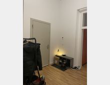 Ein Flur mit einer hellgrauen Eingangstür, hellem Holzfußboden und weiß gestrichenen Wänden. Links steht eine Kleiderstange mit Jacken, rechts befindet sich ein niedriges Regal mit Schuhen, darauf steht eine leuchtende Dekolampe; daneben liegt ein Paket.