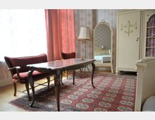 Ein klassisch eingerichtetes Zimmer mit einem dunklen Holztisch, einer roten Sitzbank und roten Vorhängen. Im Hintergrund befinden sich ein cremefarbener Kleiderschrank, ein großer Spiegel mit Verzierungen und eine Stehlampe; der Boden ist mit einem orientalischen Teppich dekoriert.