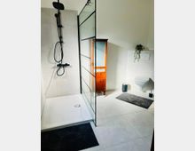 Modernes Badezimmer mit bodengleicher Dusche, schwarzer Regendusche und Glas-Duschabtrennung mit schwarzen Rahmen. Rechts sind ein wandhängendes WC mit Betätigungsplatte, ein Holzschrank sowie helle Fliesen und graue Badvorleger zu sehen.