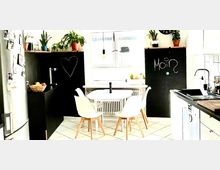 Helle Küche mit einem kleinen weißen Esstisch und vier weißen Stühlen. Schwarze Tafelflächen an den Wänden mit Kreidezeichnungen, Pflanzen auf den Schränken, ein großes Fenster in der Mitte und eine Küchentheke mit Spüle und Elektrogeräten rechts.