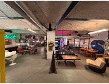 Industrieller Innenraum mit gemauerten weißen Wänden, sichtbaren Rohren und einer Kombination aus Retro- und modernen Möbeln, darunter Ess- und Loungetische sowie Sessel. Im Hintergrund befinden sich Neon-Schilder, Regale mit beleuchteten Objekten und dekorative Elemente wie eine große, blaue Skulptur in Form eines „S“.