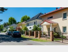 Reihen moderner Einfamilienhäuser mit unterschiedlichen, farbigen Fassaden und Ziegeldächern entlang einer ruhigen Straße; davor stehen geparkte Autos und ein schmiedeeiserner Zaun mit Backsteinpfosten umgibt die Grundstücke. Im Hintergrund sind Bäume und ein klarer blauer Himmel sichtbar.