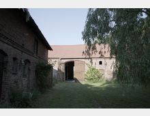 Hof mit historischen Backsteingebäuden, darunter ein einstöckiges Haus und eine Scheune mit offenen Holztoren. Der Hof ist von Gras bewachsen, und im Vordergrund hängt ein Baum mit herabhängenden Zweigen.