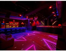 Ein düsterer Nachtclub mit roter und violetter Beleuchtung, einer langen Bar mit Hockern und Regalen voller Flaschen im Hintergrund. Die Tanzfläche ist mit gemusterten Teppichen bedeckt, und eine Discokugel hängt von der Decke, umgeben von Lautsprechern und Beleuchtungseffekten.