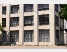 Fassade eines modernen Gebäudes mit großen rechteckigen Fenstern, die in Metallrahmen eingefasst sind. Im Vordergrund befindet sich ein leerer Parkplatz mit nummerierten Stellplätzen, teils von Bäumen gesäumt.