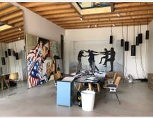 Modernes Atelier oder Büro mit offener Gestaltung. Sichtbare Holzbalkendecke, großformatige Gemälde an den Wänden, ein zentraler Schreibtisch mit Büromaterial und Stühlen sowie Pendelleuchten, die von der Decke hängen.