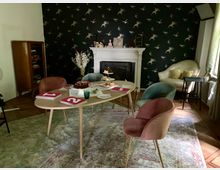 Gemütlich eingerichteter Raum mit einem ovalen Holztisch, umgeben von Stühlen in verschiedenen Farben. Im Hintergrund eine schwarze Tapete mit Vogelmuster, ein weißer Kamin mit Dekorationen sowie ein Sessel und ein Bücherregal.