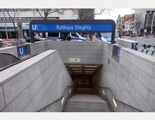 Eingang zur U-Bahn-Station „Rathaus Steglitz“ mit grauen Steinwänden, Treppen und einer Rolltreppe, die nach unten führen. Im Hintergrund sind Polizeifahrzeuge und umliegende Gebäude an einer belebten Straßenkreuzung zu sehen.