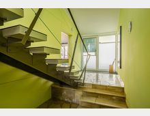 Treppenhaus mit moderner, frei schwebender Metalltreppe und Glasgeländer vor hellgrün gestrichenen Wänden. Der Raum verfügt über helle Natursteinfliesen, große bodentiefe Fenster mit Milchglas und direkten Zugang zum Außenbereich.