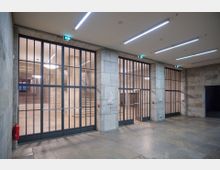 Korridor mit Steinwänden und großen Glas-Gittertüren, die einen Blick in einen dahinterliegenden Bereich mit Treppen und warm beleuchteten Pendelleuchten freigeben. An der Wand befinden sich Schilder und ein Feuerlöscher, die Umgebung ist schlicht mit Steinboden und Deckenleuchten gestaltet.