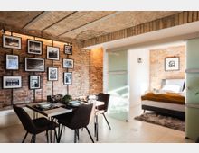 Wohnraum in einem Loft mit freiliegenden Ziegelwänden und gedecktem Esstisch vor einer Wand mit Schwarz-Weiß-Fotografien. Rechts führt eine Schiebetür aus Milchglas in ein Schlafzimmer mit weißen Wänden, Ziegelakzenten und einem Bett mit braunem Überwurf.