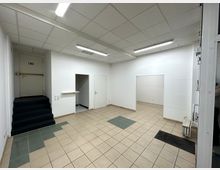 Leerer Innenraum mit weißen Wänden, beige gefliesten Böden und einigen grünen Fliesenakzenten. Links führen dunkel gepolsterte Stufen zu einer geschlossenen Tür, während seitlich offene Durchgänge erkennbar sind; die Beleuchtung besteht aus Deckenleuchtstofflampen.