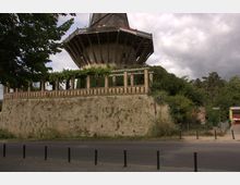 Historische Windmühle auf einem steinernen Unterbau mit umgebender Begrünung und Säulen. Im Vordergrund verläuft eine Straße mit Pollern, während Bäume und Sträucher die Umgebung ergänzen.
